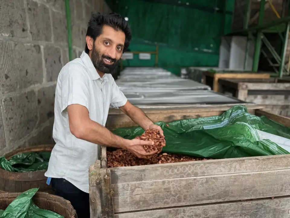 Muffadal smiling beside a wooden fermentation box of cacao, his hand on the beans