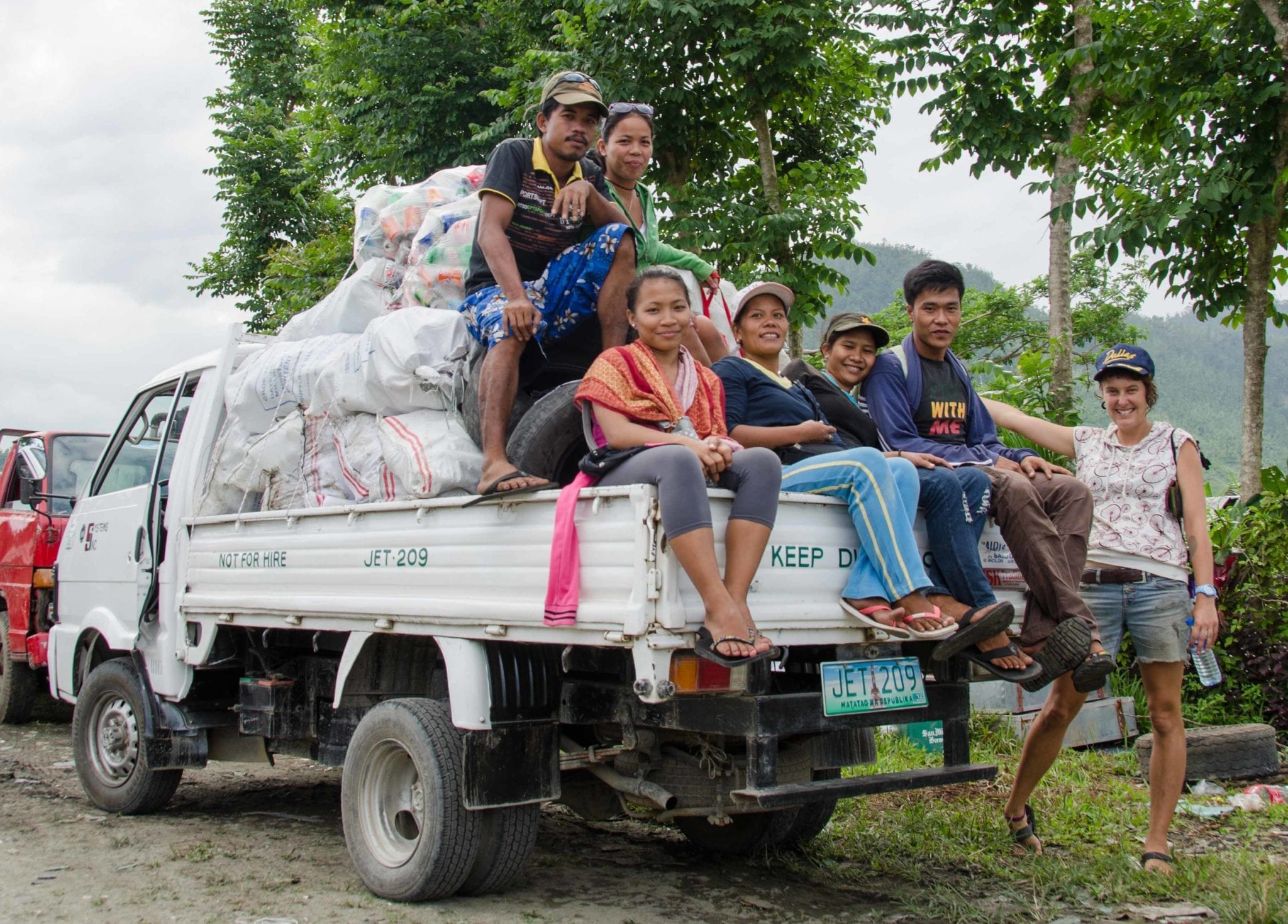Six adults sit on the back of a white pickup piled with sacks of plastic bottles, with a smiling woman standing beside it and green hills behind.